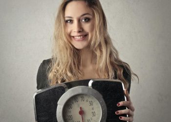 Young woman with blond hair smiling, holding a vintage bathroom scale indoors.
