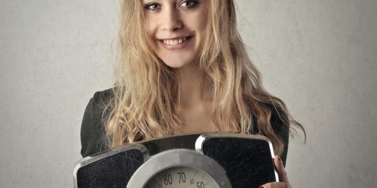 Young woman with blond hair smiling, holding a vintage bathroom scale indoors.